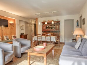 Un salon comprenant un canapé, des fauteuils, une table à manger et un coin cuisine avec des meubles en bois et des luminaires suspendus.