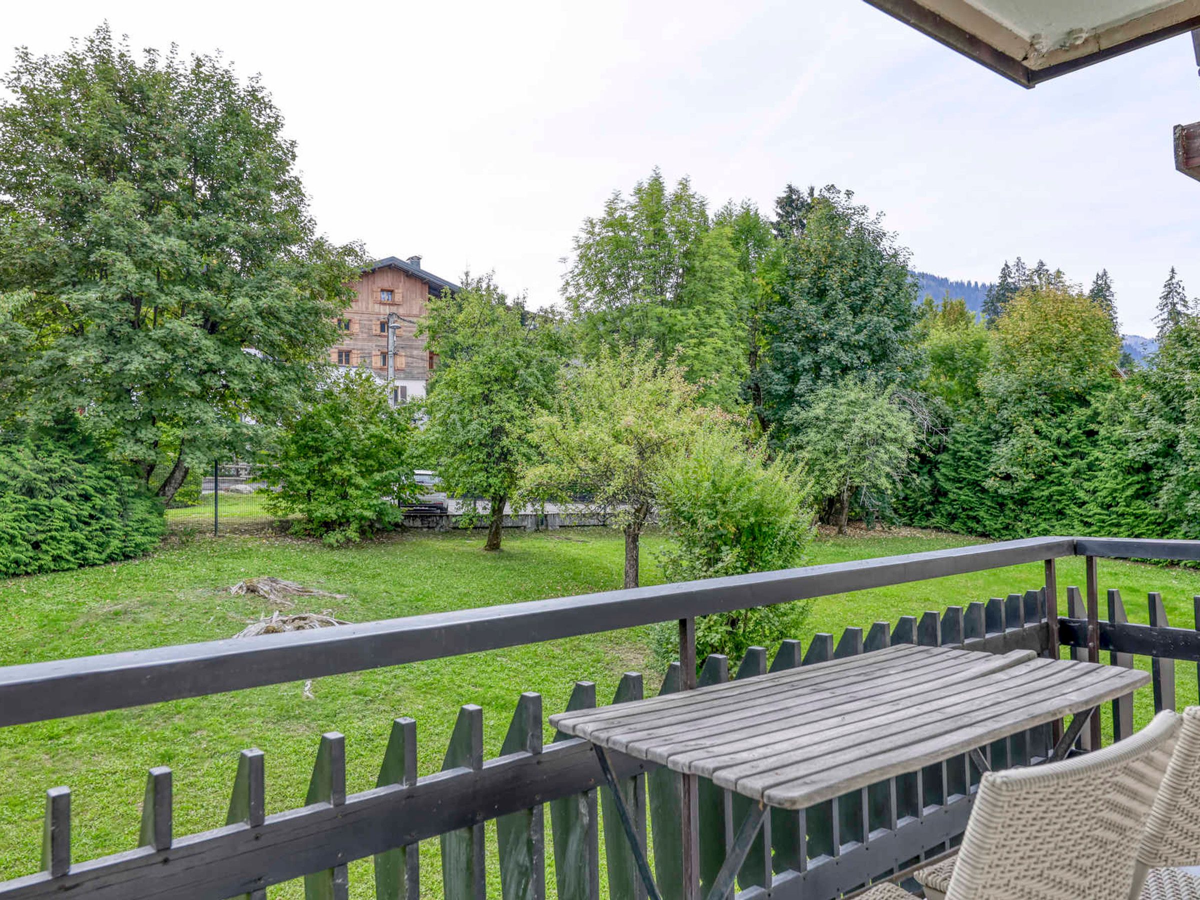 Ein Balkon mit einem Holztisch und Korbstühlen, der auf eine Grünfläche mit Bäumen und einem Gebäude im Hintergrund blickt.