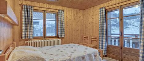 A wooden cabin bedroom with a bed, two chairs, a radiator, and large windows with curtains.