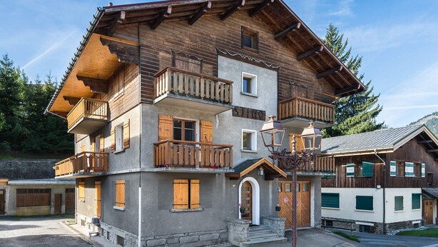Un chalet en bois à plusieurs étages, doté de balcons, d’une entrée en pierre et surplombant un ciel dégagé.