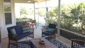 Une véranda couverte avec un ventilateur de plafond, un canapé bleu orné de coussins à rayures, deux chaises en bois, une petite table sur laquelle est posé un objet décoratif, et un tapis à motifs.