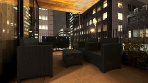 An outdoor seating area with wicker furniture, a wooden ceiling, and a view of city buildings at night. - bnb+ Kanda - Hostel (Tokyo)