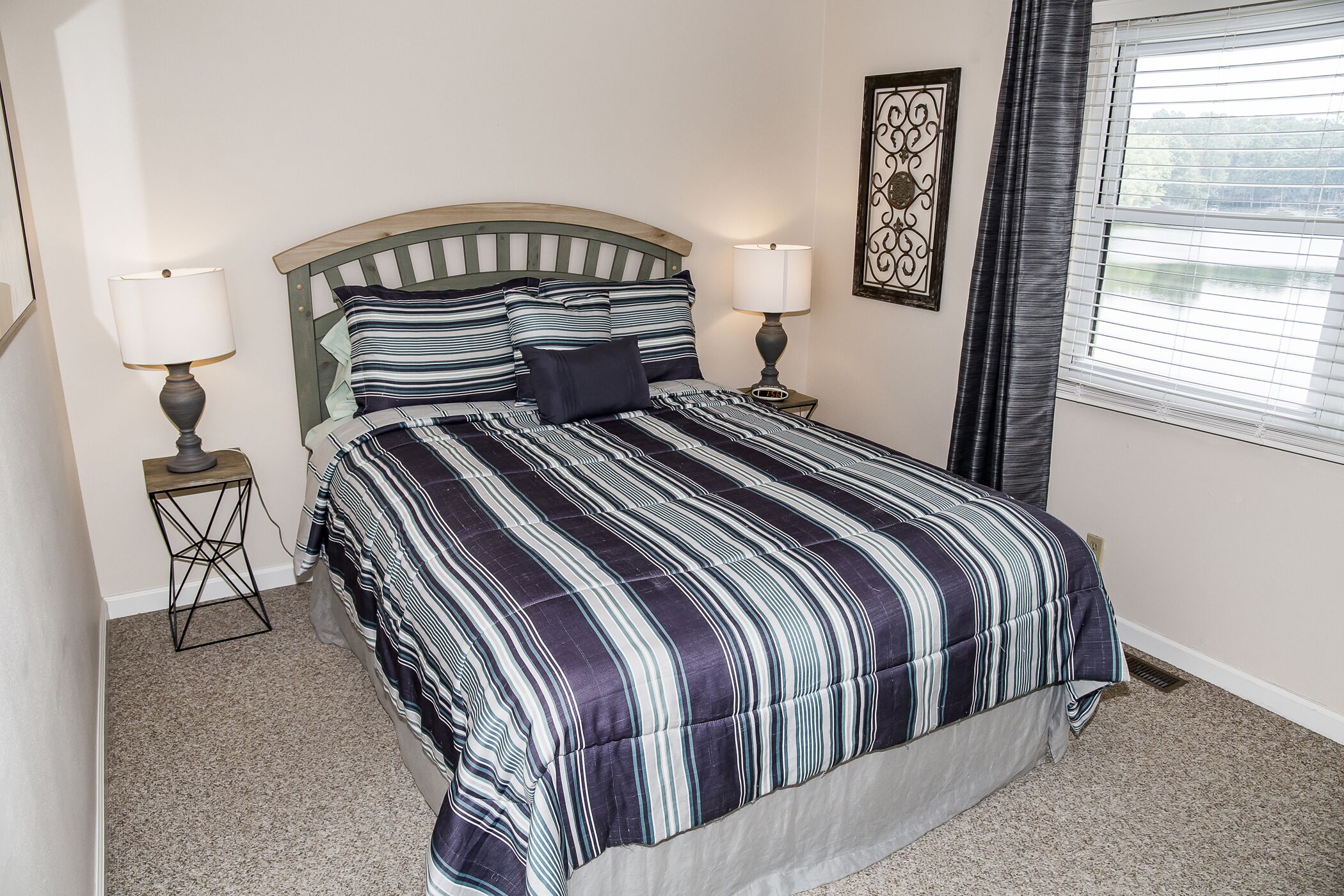 A bedroom with a bed, two bedside tables, a window with blinds, and a wall-mounted decorative piece.