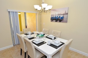 A dining area with a table set for six, white chairs, a chandelier, and a painting of boats on the wall.