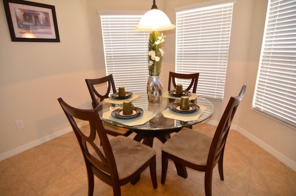 A dining area with a round glass table, four wooden chairs, a vase with flowers, and a table lamp.