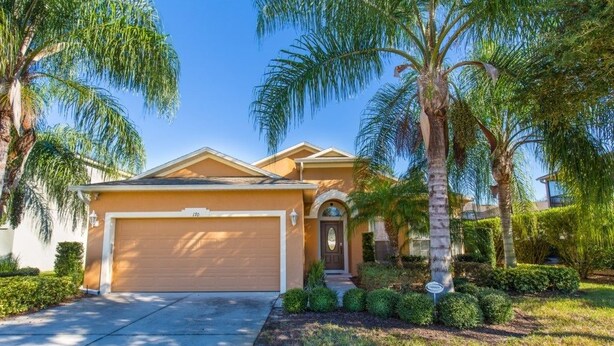 A single-story house with a beige garage door, surrounded by well-maintained landscaping including palm trees and shrubs.