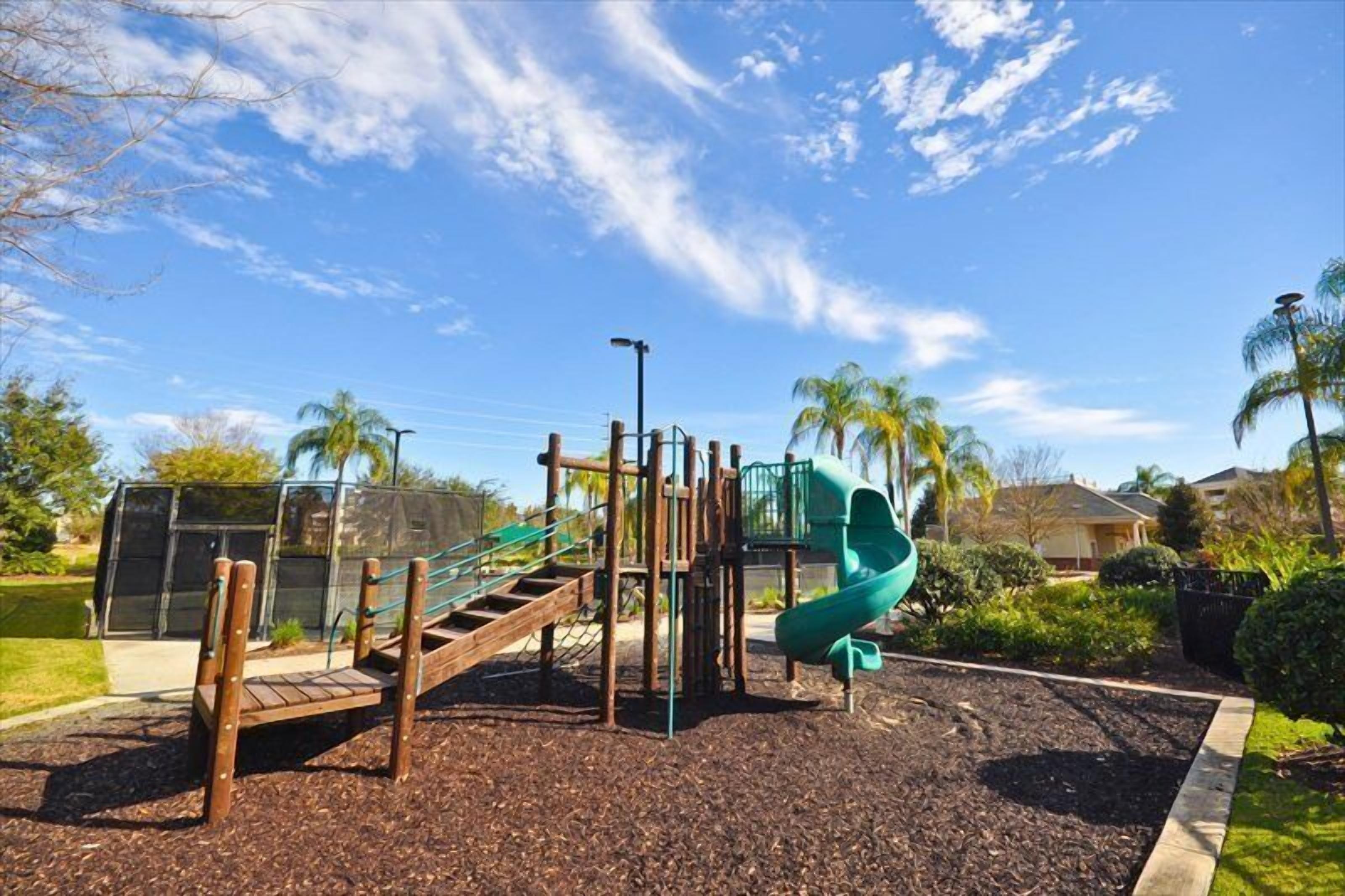 Un parque infantil con un tobogán y estructuras para escalar, rodeado de un jardín ajardinado y palmeras.