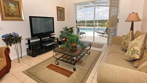 Una sala de estar con televisor, una mesa de centro con plantas, un sofá y vistas a una piscina a través de puertas de cristal grandes.
