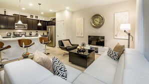 A modern living room with a white sofa, a coffee table, and a TV. There is a kitchen area with dark cabinets and a microwave. - Ev216592 - Windsor At Westside Resort - 5 Bed 4 Baths Townhome (Kissimmee)
