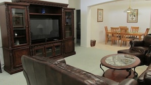 A living room with a dark wooden entertainment center, leather sofa, and a dining area with wooden chairs and a glass table. - Br52637 - Indian Creek - 5 Bed 4 Baths Villa (Kissimmee)