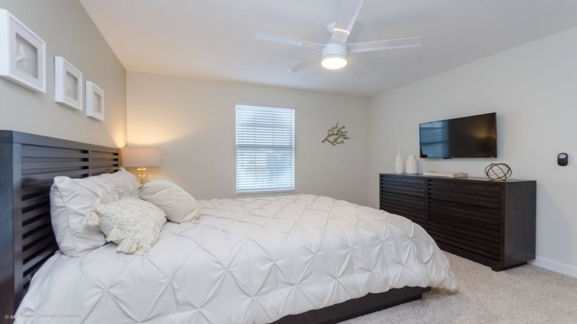 A bedroom with a bed, a ceiling fan, a TV, and a window.