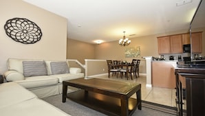 A modern living room with a sofa, coffee table, and dining area with chairs and a chandelier. - Fs55568 - Oakwater Resort - 2 Bed 2 Baths Townhome (Kissimmee)
