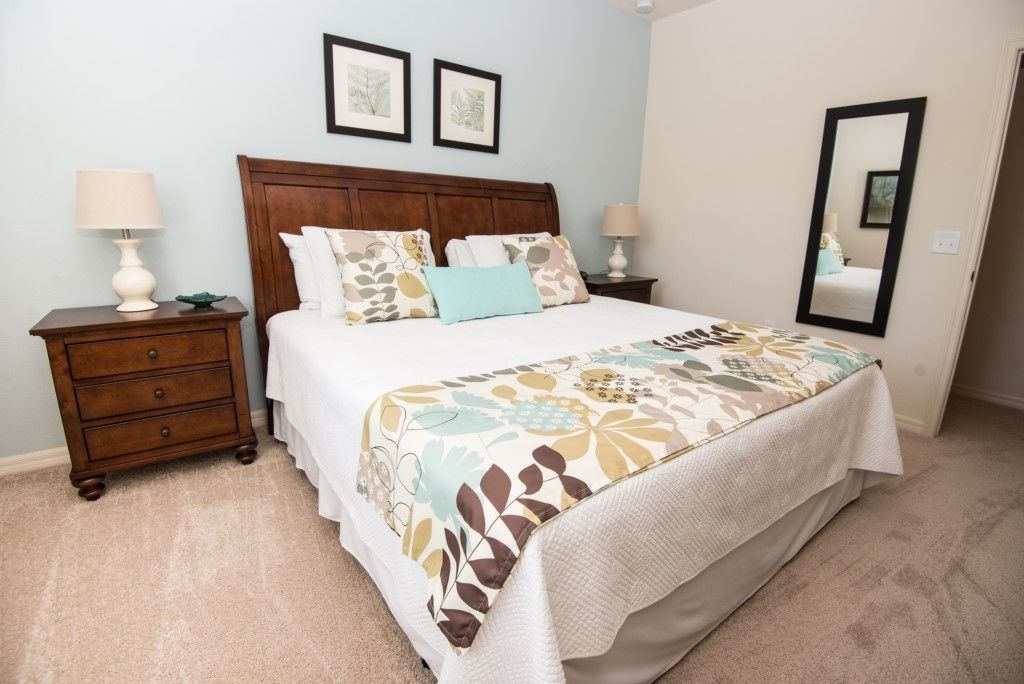 Quarto com cabeceira de madeira, cama grande com roupa de cama estampada, duas mesas de cabeceira com abajures e um espelho na parede.