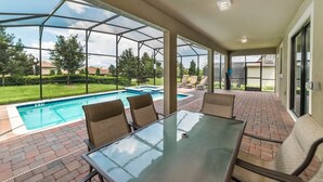 A covered patio with a glass table and chairs, a swimming pool, and a view of the surrounding area.