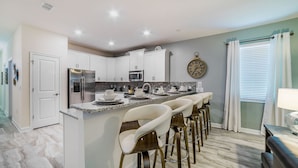 A modern kitchen with white cabinets, a stainless steel refrigerator, a microwave, and a coffee machine. There are bar stools and a clock on the wall.