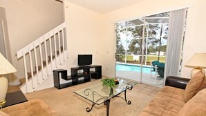 Un salon avec une table basse en verre, des canapés de couleur beige, un téléviseur et une vue sur une piscine, visible à travers de grandes portes vitrées.