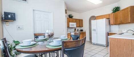 A compact kitchen with wooden cabinets, a microwave, and a refrigerator. There is a dining area with a table set for two, chairs, and a hanging light fixture.