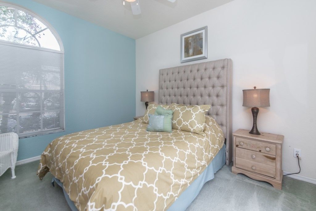 A bedroom with a tufted headboard, a bed with patterned bedding, bedside lamps, a small dresser, and a window with blinds.