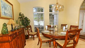 A dining area with a round table, wooden chairs, a chandelier, and a large window with blinds.