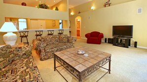 A living room with a sofa set, a coffee table, a red armchair, a TV, and a kitchen area in the background.