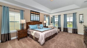 Une chambre spacieuse avec un grand lit, deux tables de chevet, une petite table et une commode. Elle est dotée de plusieurs fenêtres avec des rideaux, offre une vue sur l’extérieur et possède un luminaire au plafond.
