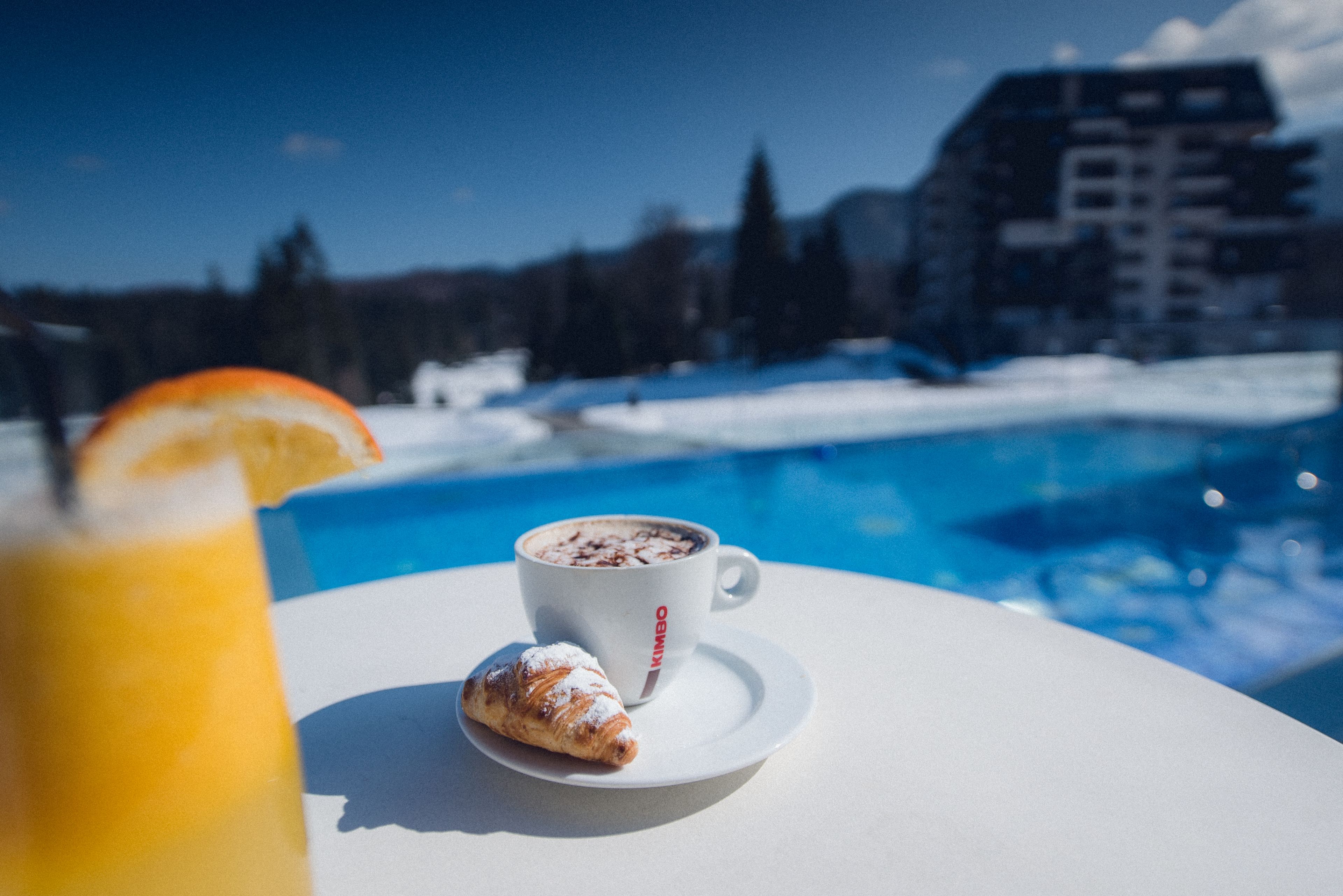 A white table with a cup of coffee and a pastry, overlooking a swimming pool and a snowy landscape.