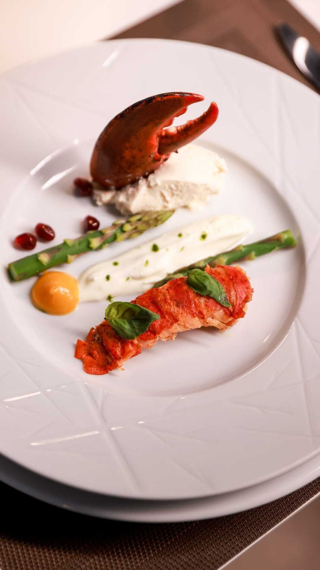 Ein weißer Teller mit einem Krabben, Spargel, einem kleinen Ei und einem Stück Fleisch, garniert mit Basilikum.