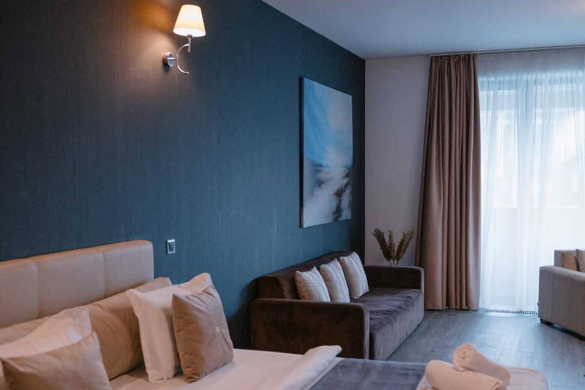 Habitación de hotel con una cama grande, un sofá más pequeño, un escritorio y una silla. Se observa un cuadro en la pared y una ventana con cortinas.