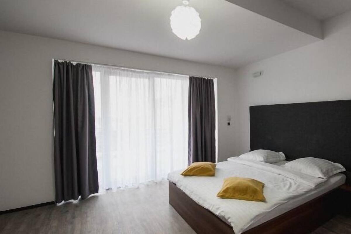 Un dormitorio con cama, dos almohadas, cabecera y ventana con cortinas.