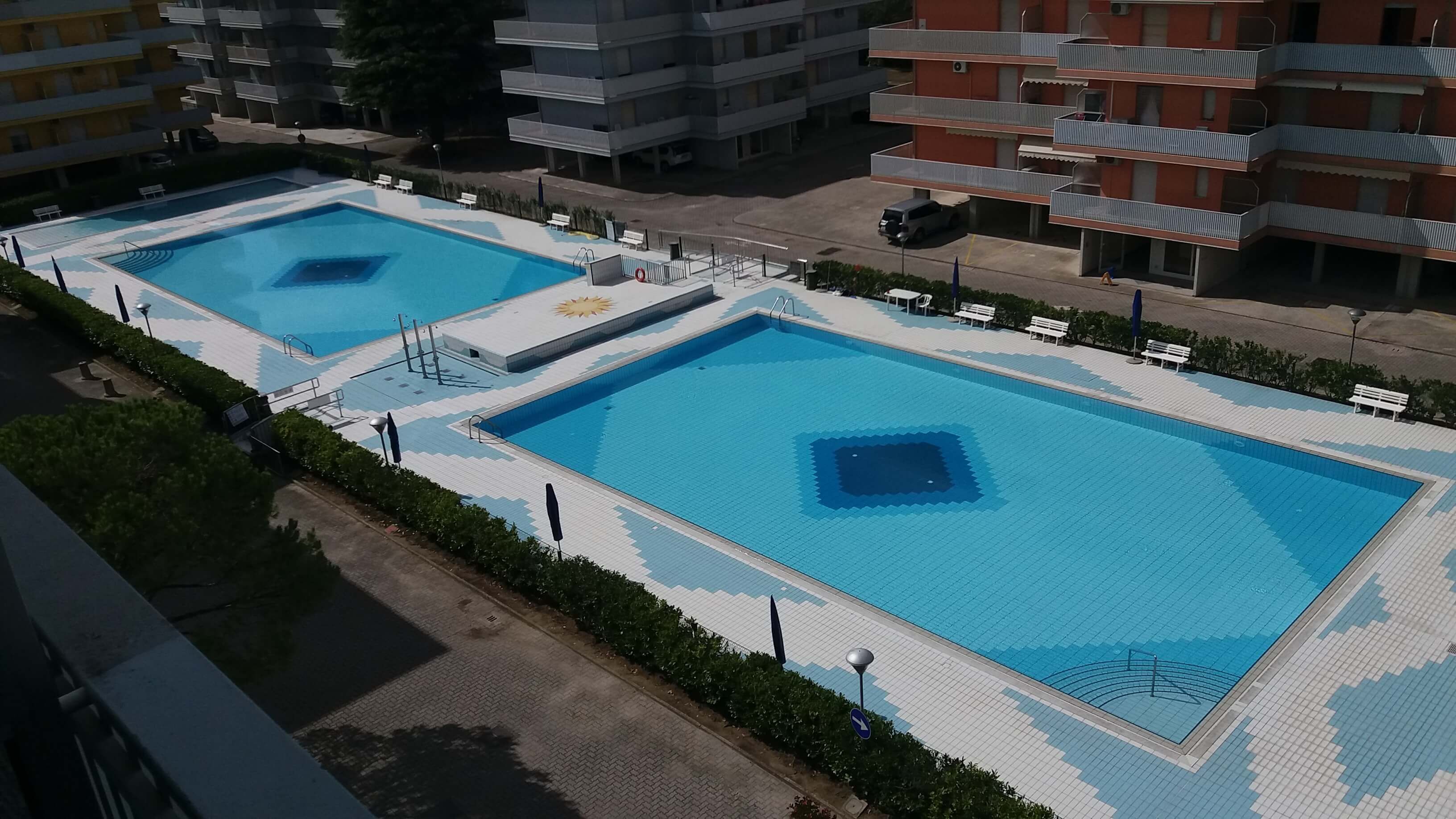 Una amplia piscina al aire libre con dos zonas separadas, rodeada por un sendero pavimentado y bancos.