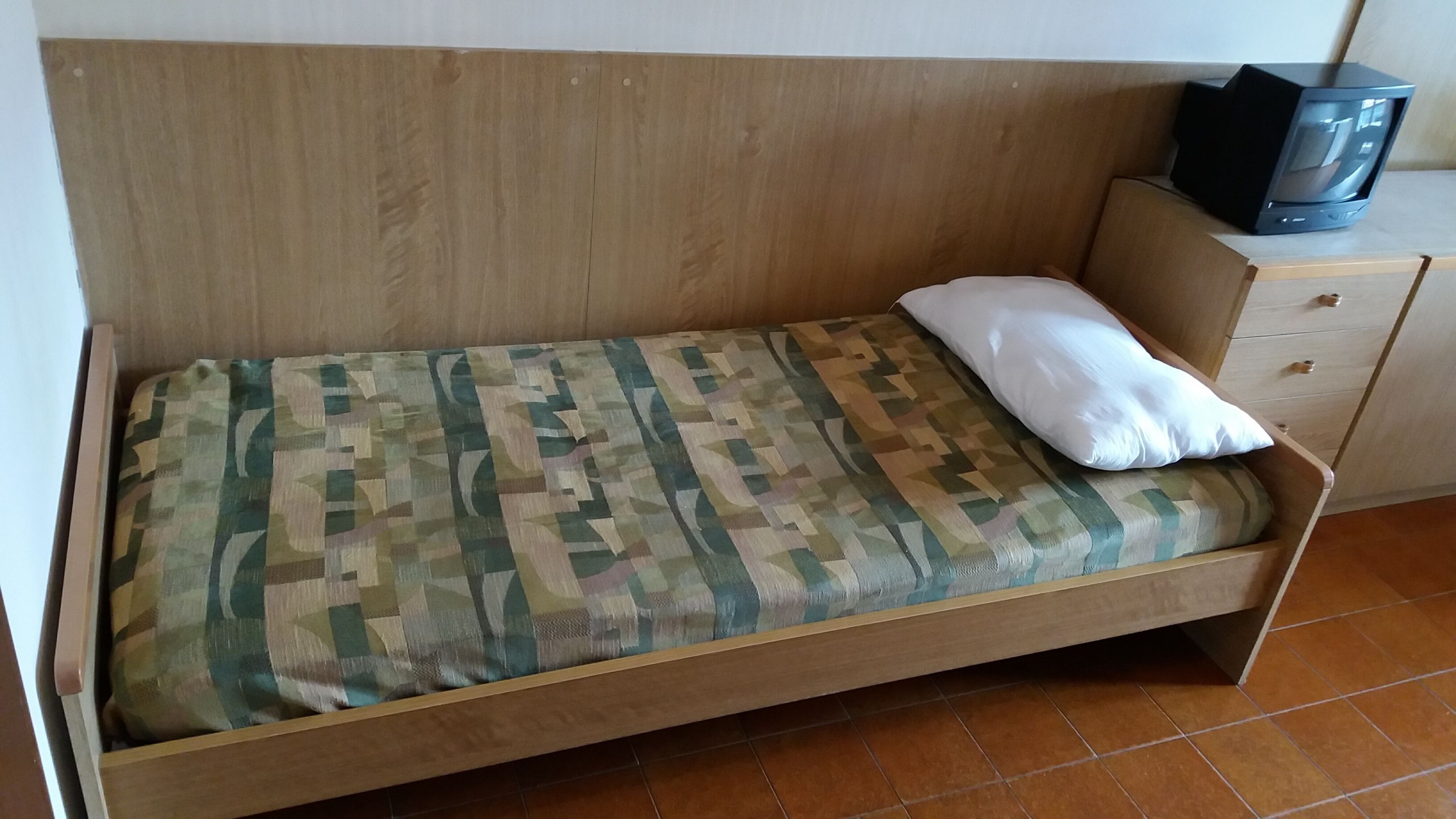 Un letto singolo con un materasso a fantasia camouflage, un cuscino bianco e una testiera in legno. Una piccola comodino in legno con una televisione si trova a destra del letto.