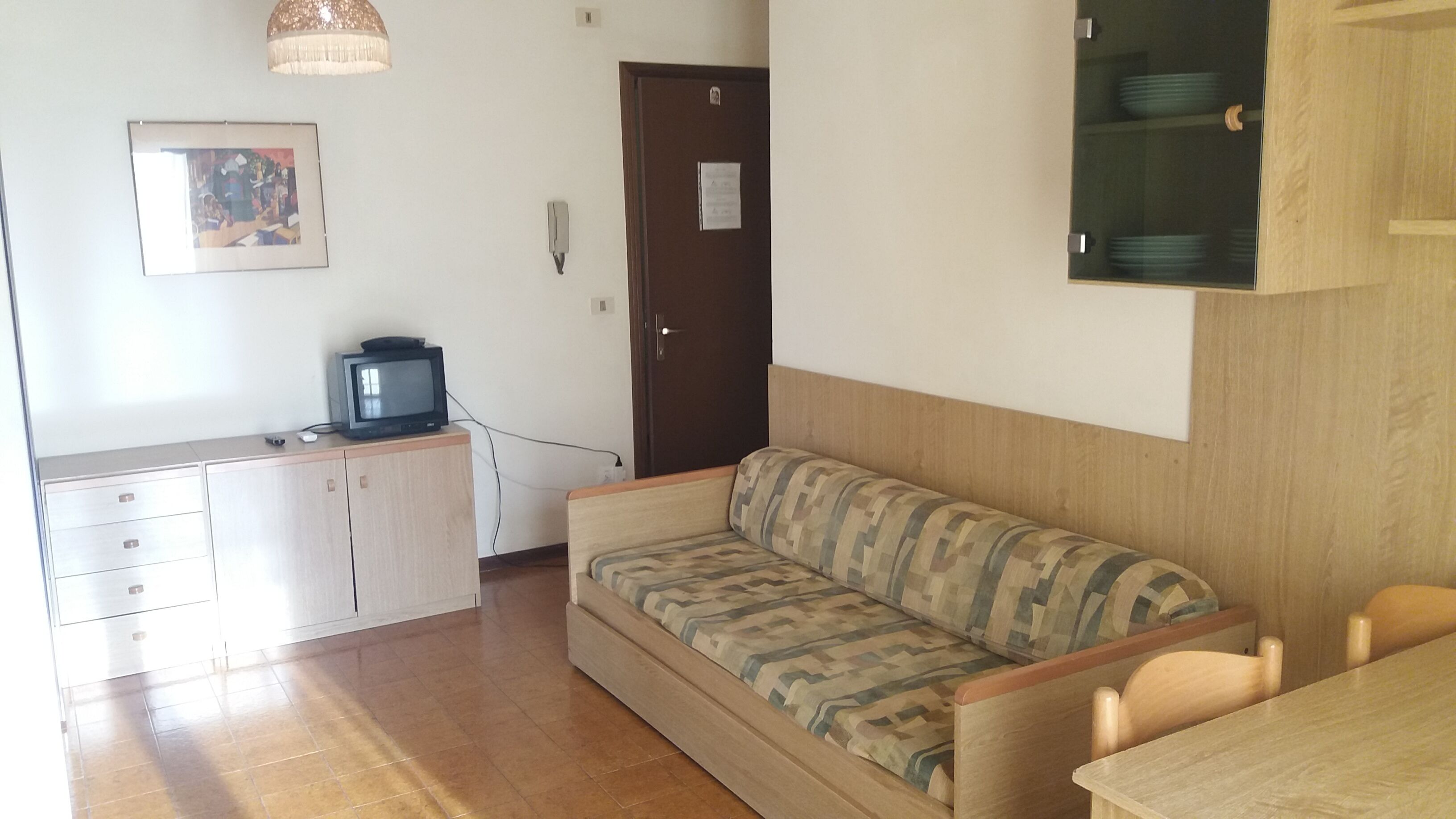 A small, well-lit room with a wooden floor, a sofa, a small table, a cabinet, and a television on a stand.