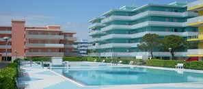 A swimming pool area with a tiled deck, surrounded by greenery and modern buildings with balconies. - Residence Valbella (San Michele al Tagliamento)