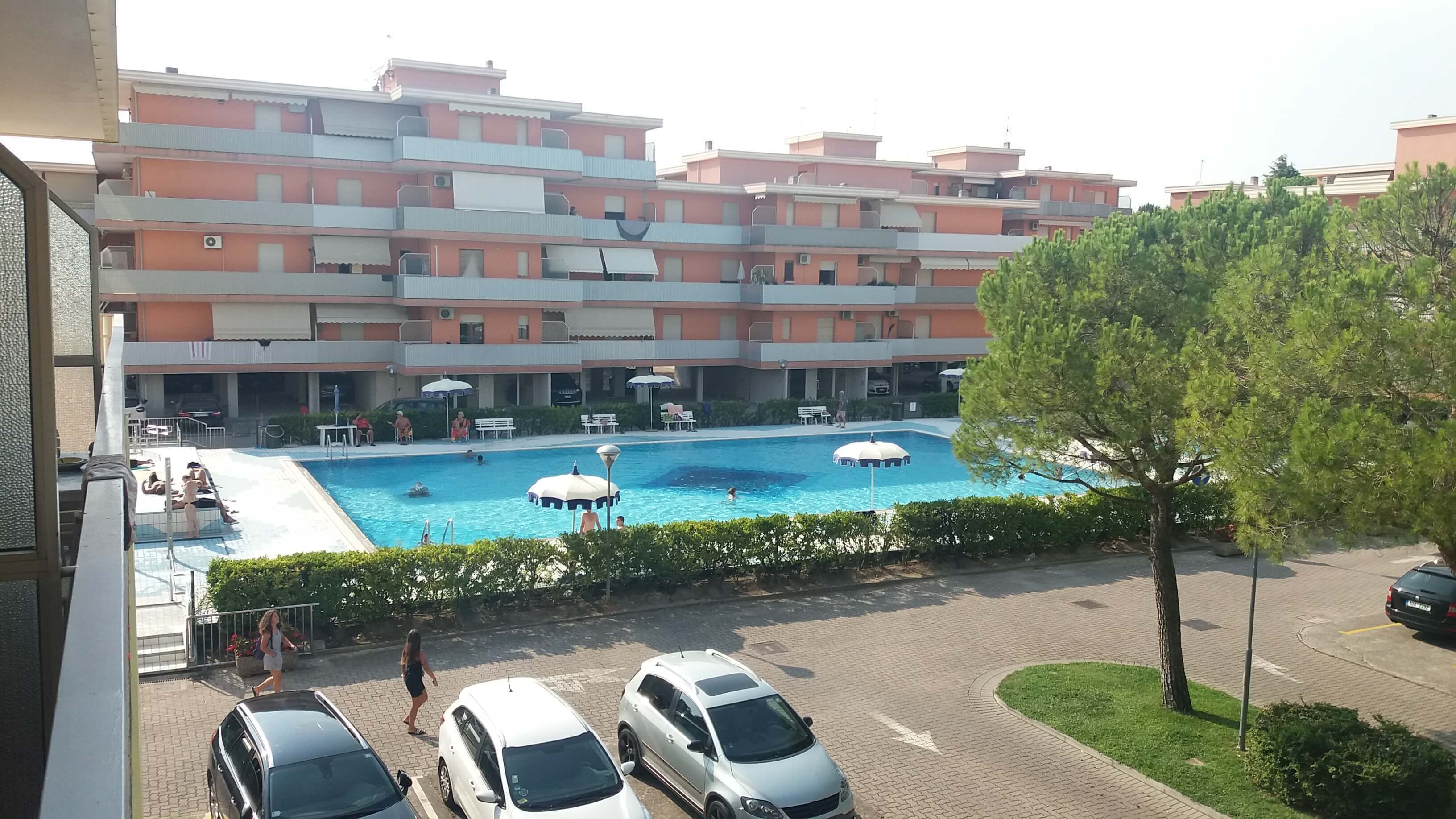 A swimming pool with lounge chairs and umbrellas, surrounded by a parking area and a multi-story building with balconies.