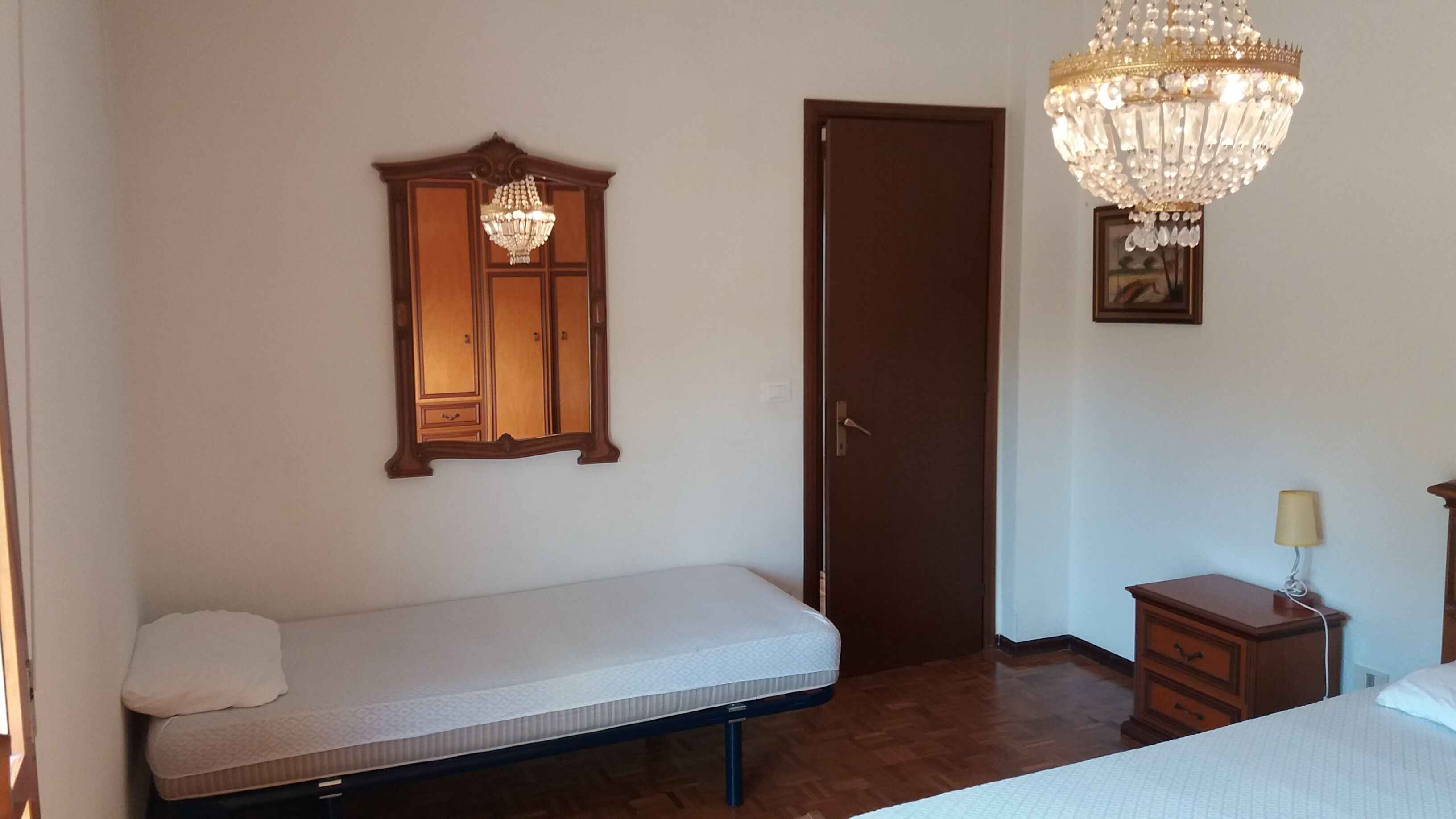 Un dormitorio con una cama individual, un armario de madera, una puerta, una araña y una mesita de noche con lámpara.
