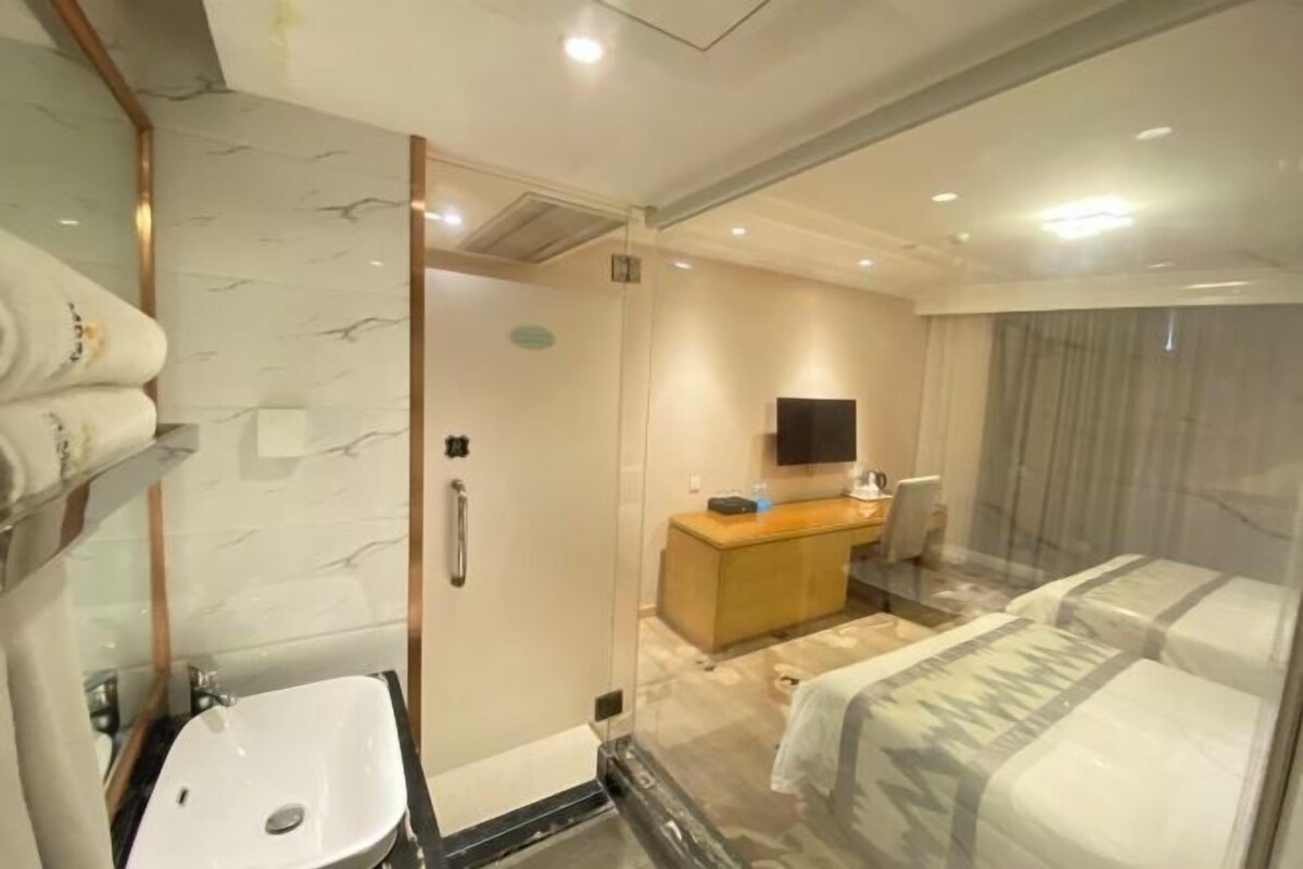 A hotel room with a glass shower enclosure, a sink, a wooden vanity, a bed, and a television.