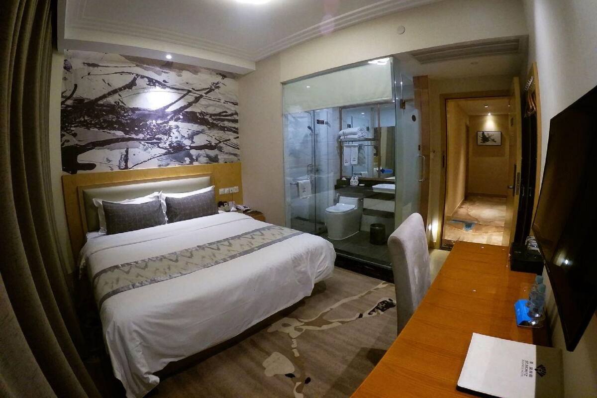 A hotel room with a bed, a desk, a chair, a bathroom with a glass partition, and a television.