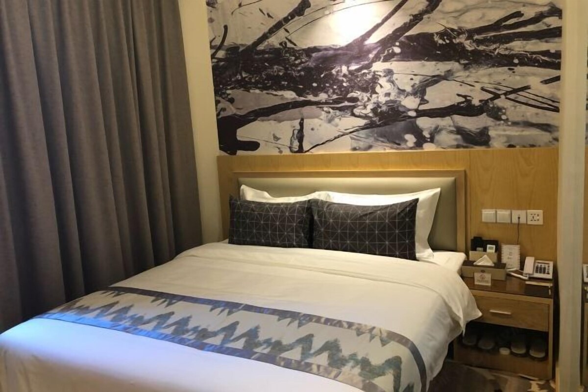 A hotel room with a bed, a nightstand, a shoe rack, and a wall with a black and white abstract painting.