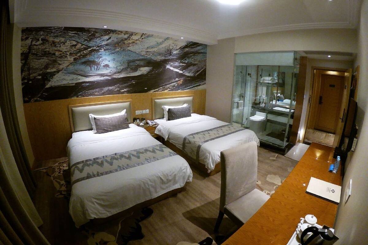 A hotel room with two beds, a large mural on the wall, a glass-enclosed bathroom, and a wooden desk with a kettle.
