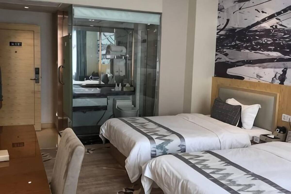 A hotel room with two beds, a desk, a chair, a glass door leading to a bathroom, and a large abstract painting on the wall.