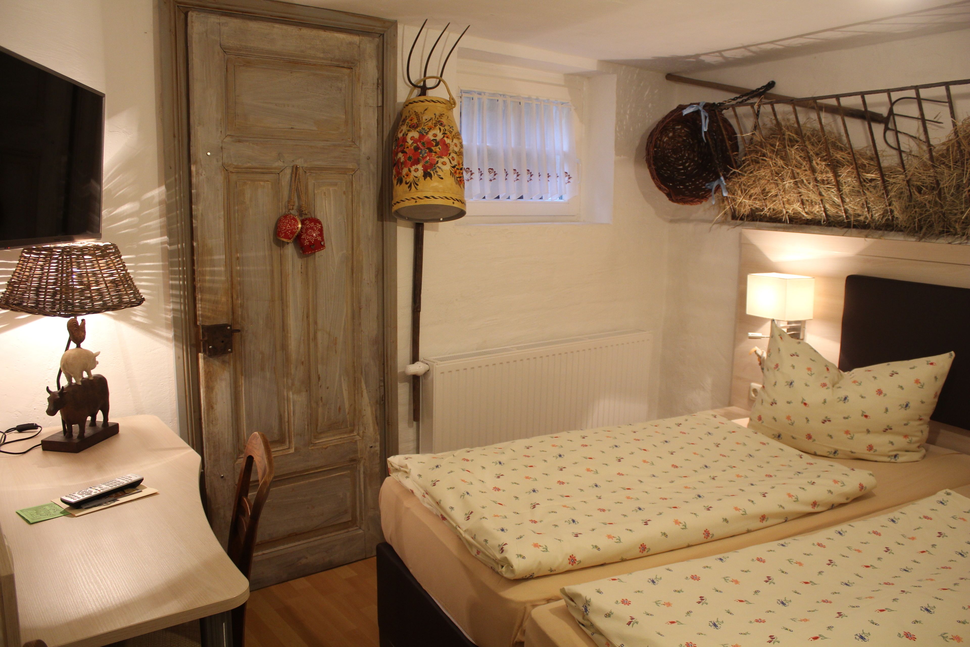 A room with two beds, a table with a lamp, a radiator, and a door with a decorative item.