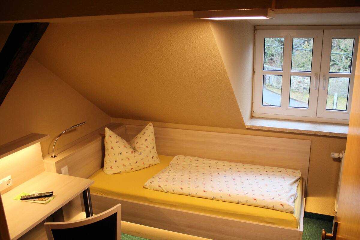 Una habitación pequeña con cama individual, escritorio, ventana y ducha.