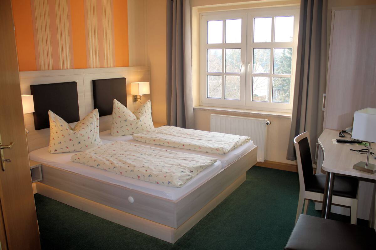 Una cama doble con dos mesitas de noche, una ventana con cortinas, un escritorio con una silla y un radiador.