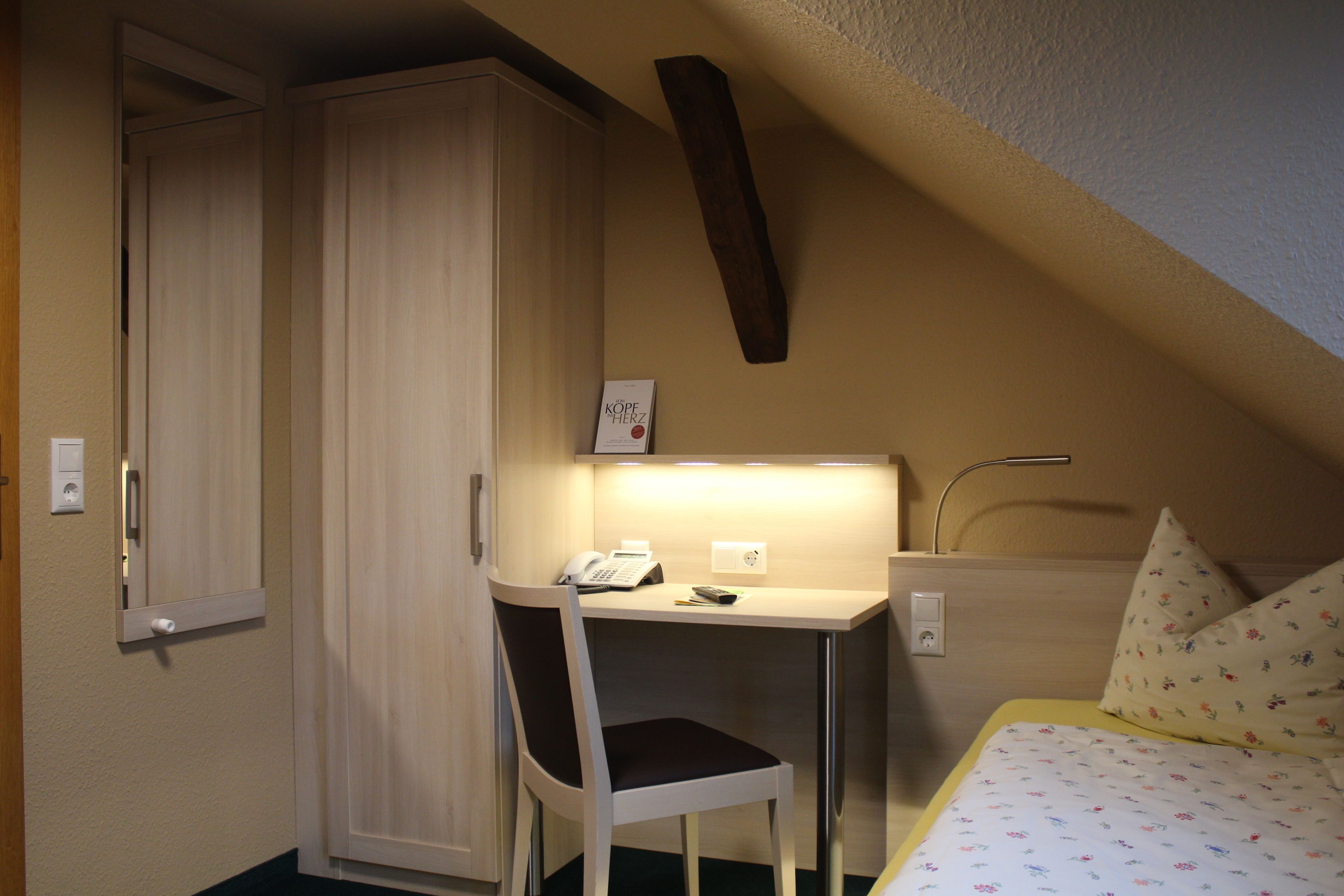 Une petite chambre à coucher avec un lit, un bureau et une chaise. On y trouve une armoire et un miroir accroché au mur.