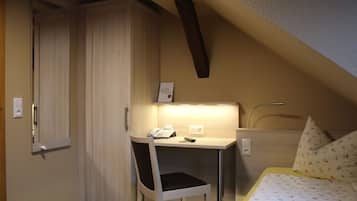 Une petite chambre à coucher avec un lit, un bureau et une chaise. On y trouve une armoire et un miroir accroché au mur.