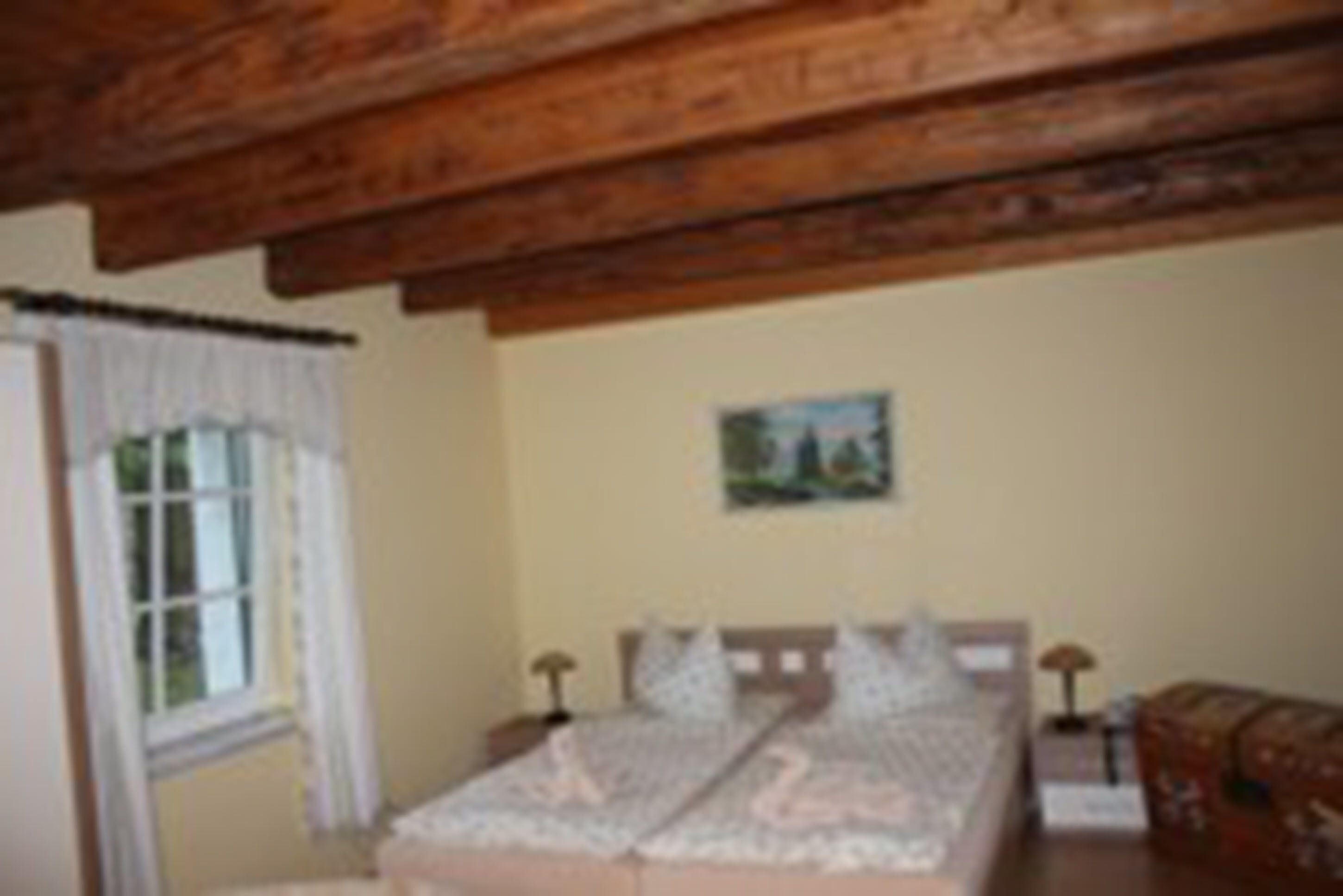A bedroom with a wooden ceiling, a bed with a white and pink bedspread, a nightstand with a lamp, and a window with curtains.