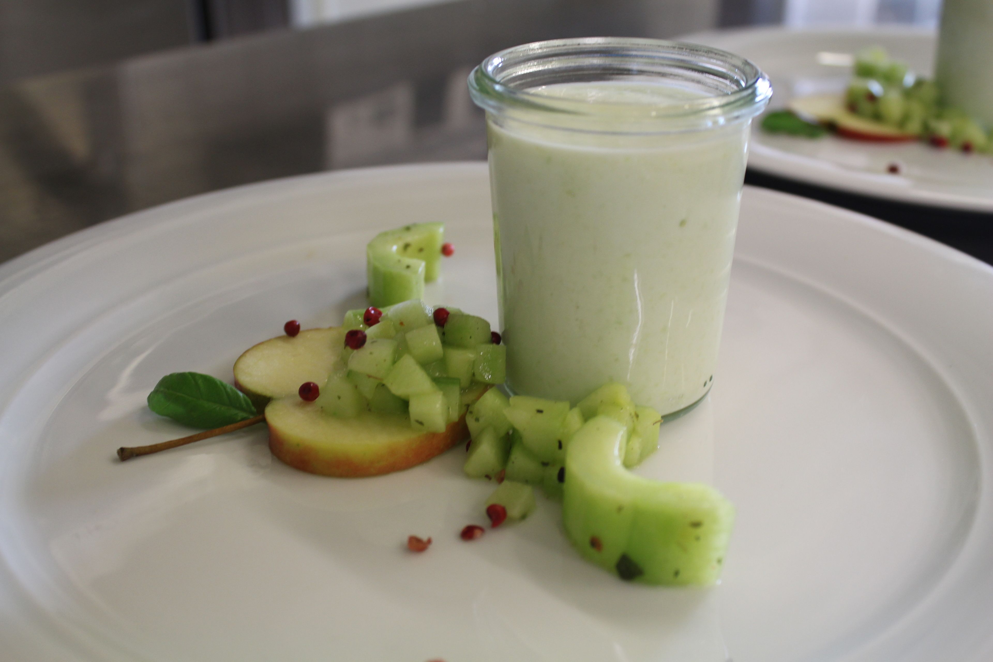 Une assiette blanche avec un verre de smoothie vert, des tranches de pomme et une feuille de menthe.