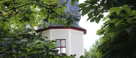 Un pequeño edificio blanco con techo rojo y una ventana, rodeado de abundante follaje verde.