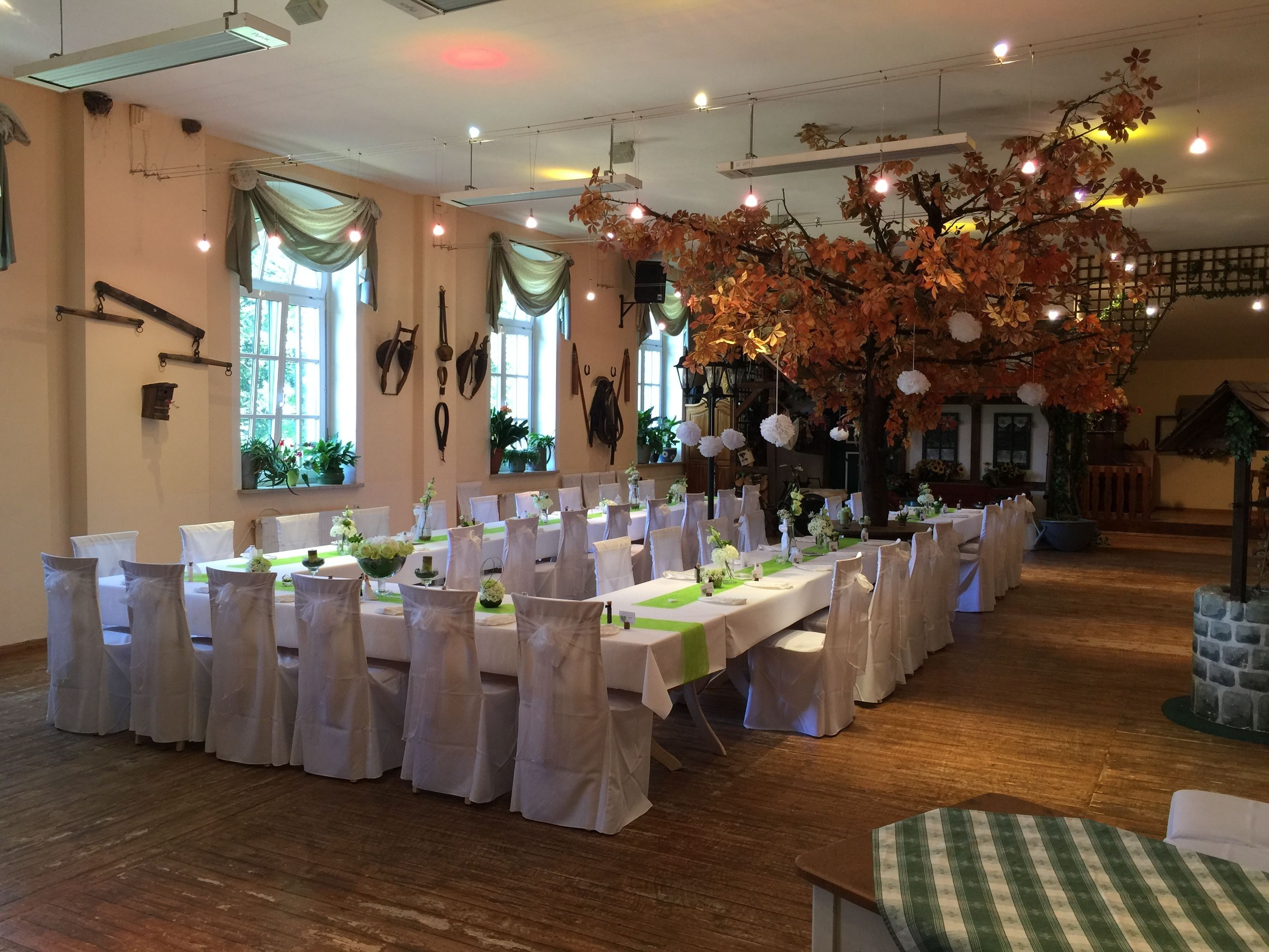 A banquet hall with long tables set for a formal event, featuring white tablecloths and chairs, and a large artificial tree with autumn leaves.