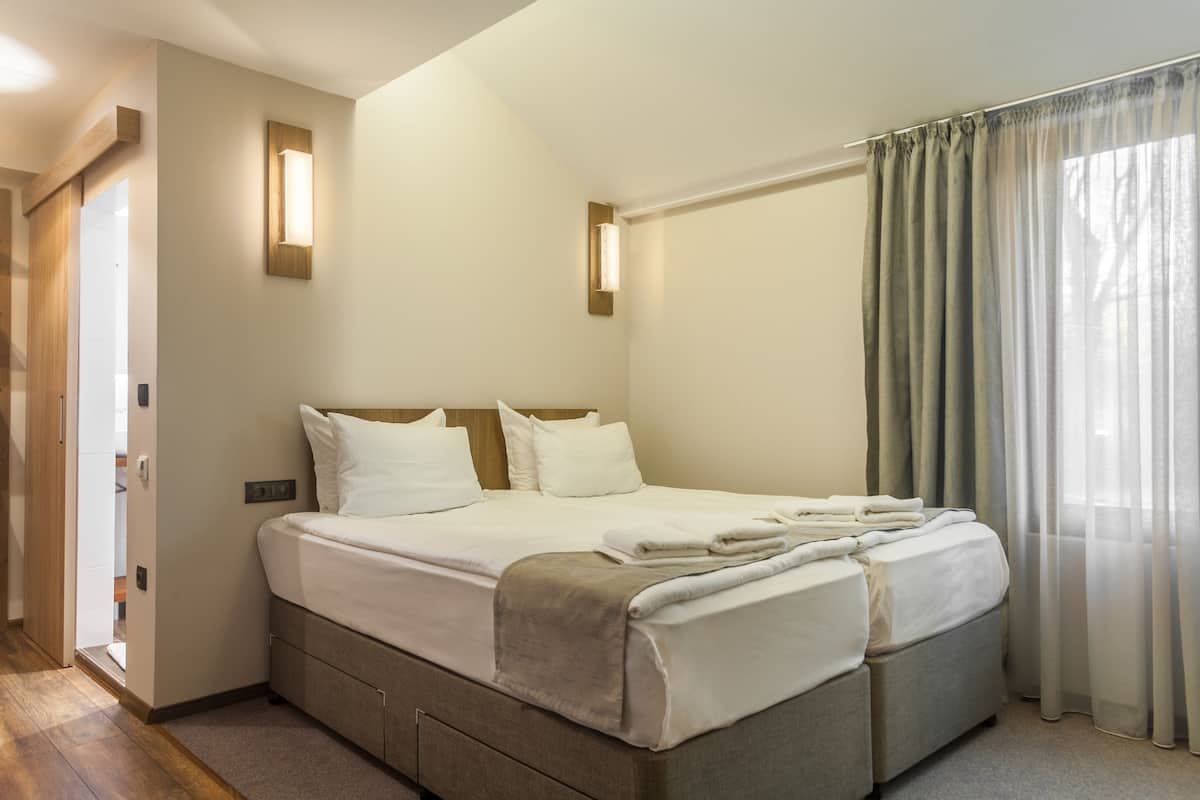 Quarto de hotel moderno com uma cama grande, mesa de cabeceira, luminária na parede e janela com cortinas.